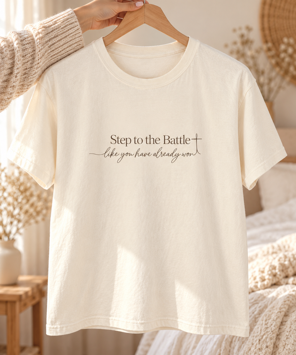 An off-white short-sleeve t-shirt with a printed motivational quote on the chest, hanging on a wooden hanger held by a person wearing a beige knit sweater in a cozy bedroom setting | Faithful Finds