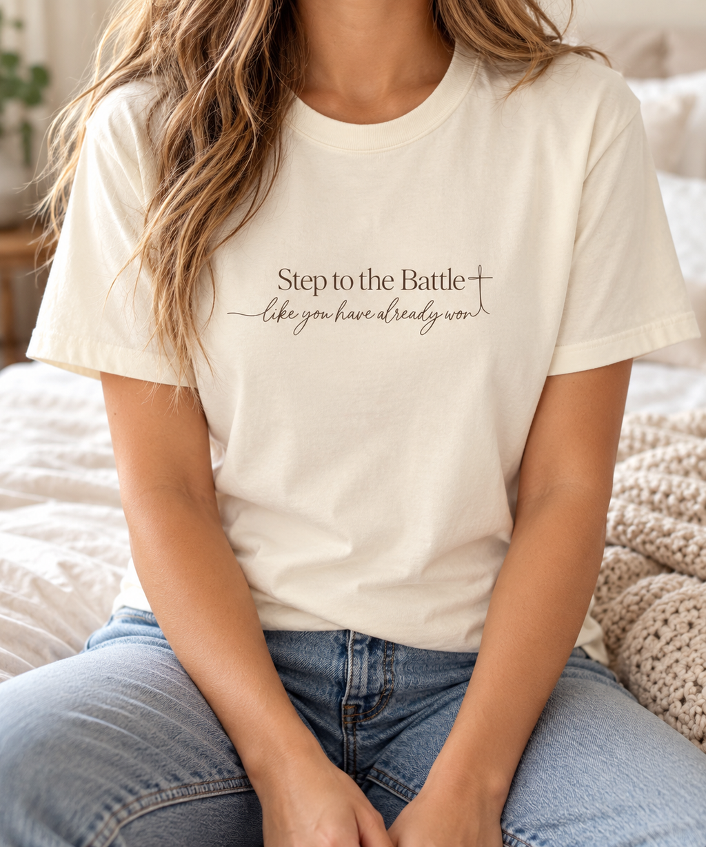 A woman wearing a cream-colored short-sleeve t-shirt with the text 'Step to the Battle like you have already won' printed in brown on the chest, paired with blue jeans in a cozy bedroom setting | Faithful Finds