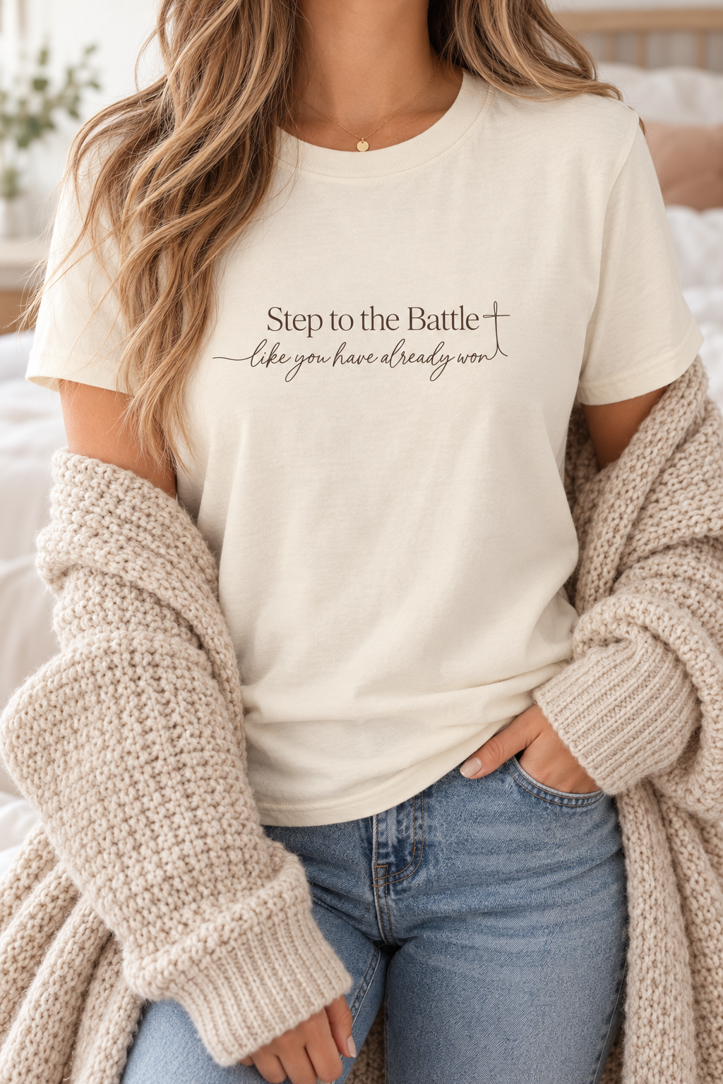 A woman wearing a cream-colored graphic t-shirt with the text 'Step to the Battle like you have already won' and a cross symbol, styled with a chunky beige knit cardigan and blue jeans | Faithful Finds