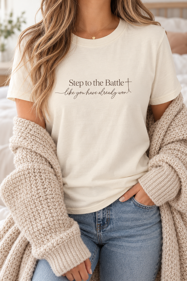 A woman wearing a cream-colored graphic t-shirt with the text 'Step to the Battle like you have already won' and a cross symbol, styled with a chunky beige knit cardigan and blue jeans | Faithful Finds