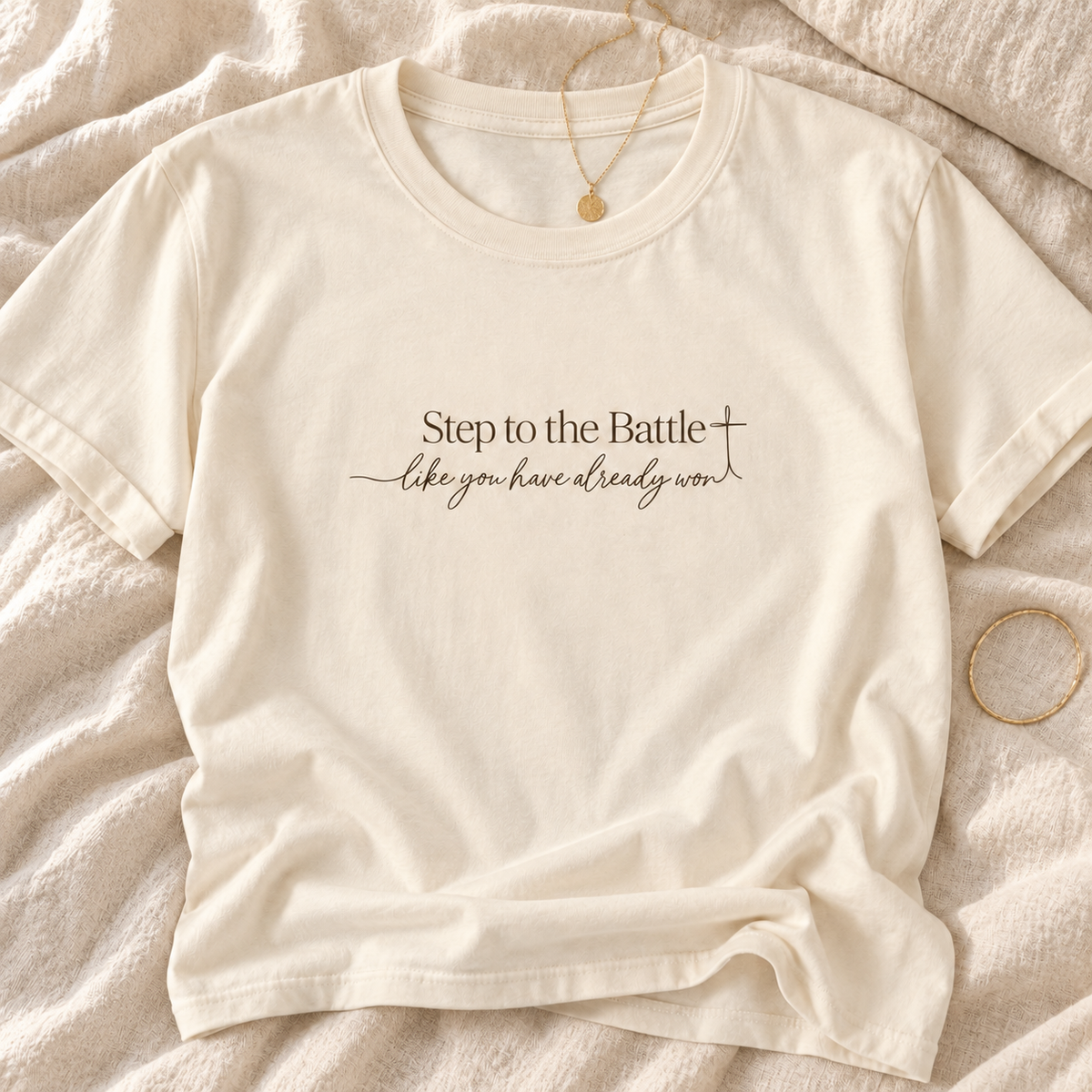 An off-white short-sleeve crewneck t-shirt with the text 'Step to the Battle like you have already won' printed in dark brown on the chest, styled flat on a beige textured blanket with a gold necklace and bracelet | Faithful Finds