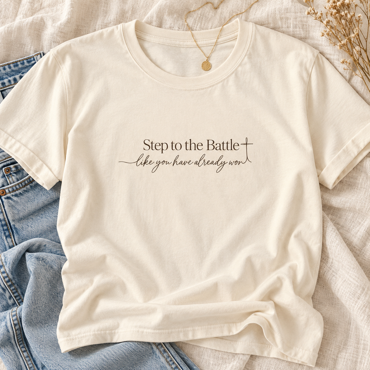 An off-white short-sleeve t-shirt with the printed text 'Step to the Battle like you have already won' and a small cross, styled flat-lay on blue jeans and a beige linen fabric background | Faithful Fins