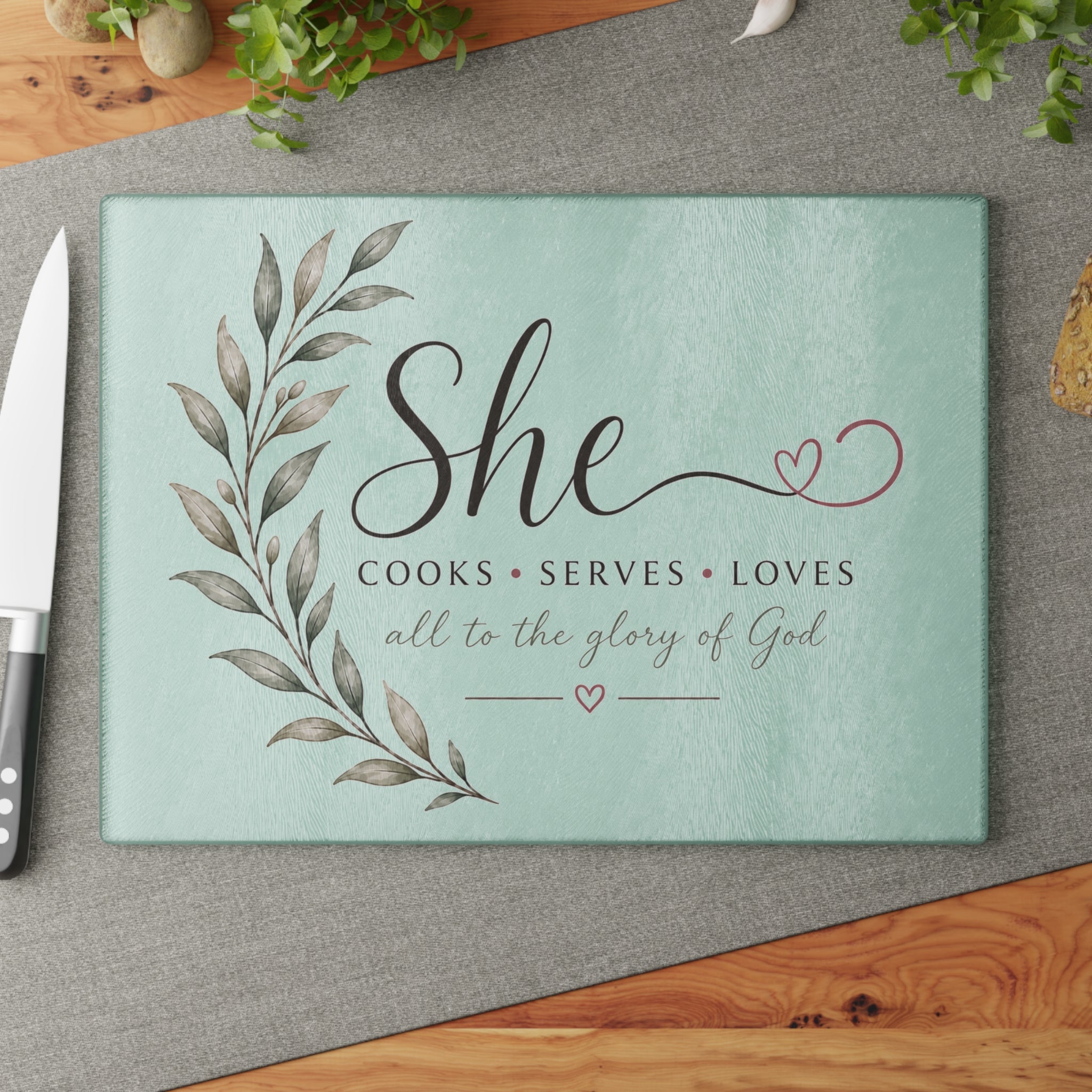 Tempered glass cutting board with 'She Cooks Serves Loves — All to the Glory of God' script on a mint background.